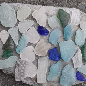 Puede incluir: Una colección de piezas de vidrio de mar de varias formas y tamaños, incluyendo tonos claros, azules y verdes, dispuestas sobre una piedra gris. El vidrio de mar está esparcido sobre la piedra, creando una estética natural inspirada en la playa.
