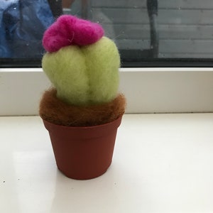 May include: A small felt cactus in a brown pot with a pink flower on top. The cactus is green and the pot is brown.