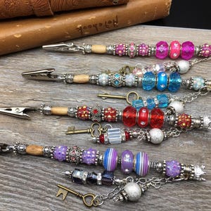 May include: Four beaded pen holders with clips. Each pen holder has a wooden stick with beads, charms, and a key attached. The beads are in various colors, including pink, blue, red, and purple.