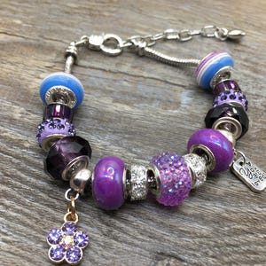 Puede incluir: Pulsera de abalorios morada y azul con un abalorio de flor y un abalorio de plata que dice "You are loved".