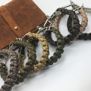 May include: A collection of braided paracord bracelets in various camouflage patterns, including green, brown, and tan. Each bracelet features a silver-colored metal clasp. A brown leather wallet with an embossed design is in the background.