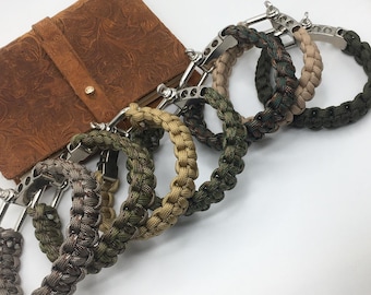 May include: A collection of braided paracord bracelets in various camouflage patterns, including green, brown, and tan. Each bracelet features a silver-colored metal clasp. A brown leather wallet with an embossed design is in the background.