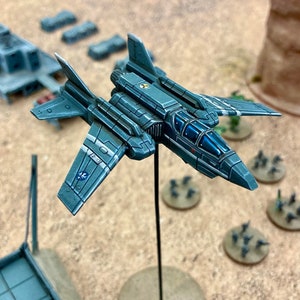 Könnte beinhalten: Ein graues und blaues Miniatur-Raumschiff mit einem klaren Cockpit, montiert auf einem schwarzen Ständer. Das Raumschiff hat ein detailliertes Design mit weißen Streifen an den Flügeln.