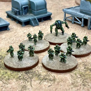 May include: A collection of miniature wargaming figures featuring small soldiers in green armour and a single mechanical walker unit. Each figure is mounted on a circular, textured base painted to resemble sand, arranged in small tactical groups.