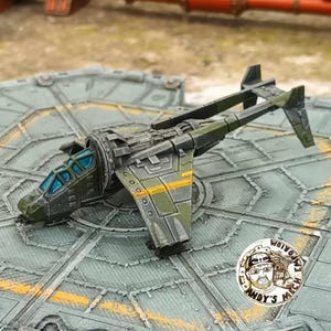 Puede incluir: Un modelo de avión en miniatura de color verde oliva con diseño de doble cola, que presenta una cabina con ventanas azul brillante, detalles mecánicos grises y una franja amarilla en las alas, sobre una plataforma de aterrizaje gris texturizada.