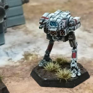 May include: A miniature bipedal robot figurine painted in white with red accents and a blue cockpit window. The mechanical figure stands on a black hexagonal base featuring small tufts of green grass, showcasing detailed paneling and weapon ports.