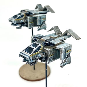 Könnte beinhalten: Zwei graue und weiße Miniatur-Raumschiffe mit gelben Akzenten. Die Raumschiffe haben ein futuristisches Design mit mehreren Fenstern und einem detaillierten Äußeren. Die Raumschiffe stehen auf schwarzen Ständern.