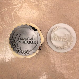 Peut inclure: Un cupcake avec un décor comestible et un tampon assorti. Le décor rond porte l'inscription "Umrah MUBARAK" dans un motif floral. Le tampon blanc présente le même motif, idéal pour les décorations.