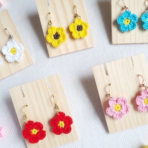 May include: A set of six pairs of crocheted flower earrings. Each pair features a different color flower, including yellow, white, blue, pink, and red.