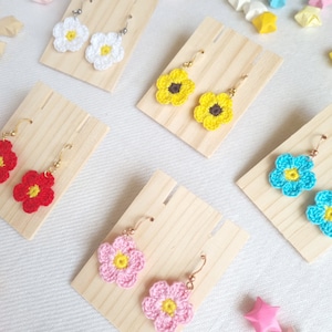 May include: Five pairs of crocheted flower earrings displayed on wooden earring stands. The earrings are in various colors, including yellow, red, pink, white, and blue.