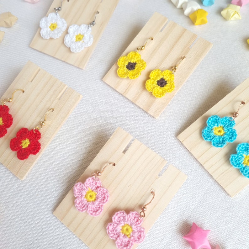 May include: Five pairs of crocheted flower earrings displayed on wooden earring stands. The earrings are in various colors, including yellow, red, pink, white, and blue.