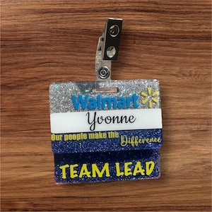 May include: A rectangular glittery badge reel featuring the blue Walmart logo, the name Yvonne in black script, and the yellow text Our people make the Difference and TEAM LEAD. The badge is attached to a silver metal clip at the top.