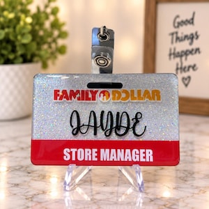 May include: A rectangular badge holder featuring a silver glitter background with the Family Dollar logo at the top. The name JAYDE is written in black script in the center, and a red horizontal bar at the bottom displays STORE MANAGER in white.