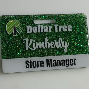 May include: A rectangular name badge with a green glitter background. The badge features the Dollar Tree logo, the name "Kimberly", and the title "Store Manager" in white and black text.