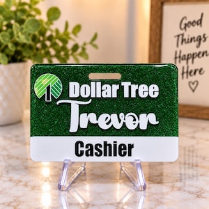May include: A rectangular name badge with a dark green glitter finish. It features the Dollar Tree logo, the name Trevor in white script, and the word Cashier in black text on a white background. The badge has a horizontal slot at the top.