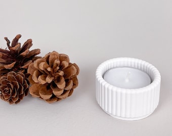 May include: White ceramic candle holder with a fluted design. The holder is empty and sits on a white surface next to three brown pine cones.
