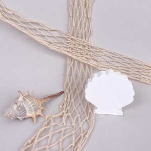 Könnte beinhalten: Ein weißes Muschel-förmiges Ornament mit einem Hintergrund aus einem Seilnetz. Die Muschel ist aus Holz und hat eine glatte Oberfläche. Das Seilnetz ist aus Naturfasern und hat einen rustikalen Look.