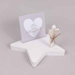Könnte beinhalten: Ein weißer sternförmiger Holzständer mit einer hellvioletten Karte, auf der "Happy Birthday, mein Lieblingsmensch" in einem goldenen Herz steht. Ein kleiner Strauß getrockneter Blumen und eine weiße Kerze stehen auf dem Ständer.