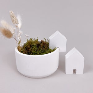 Könnte beinhalten: Ein weißer Keramiktopf mit einem moosbewachsenen Oberteil und zwei kleinen weißen Häusern an der Seite. Der Topf ist mit getrockneten weißen Blumen gefüllt.