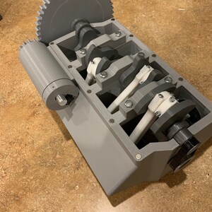 以下が含まれることがあります： クランクシャフトとコンロッドを備えた、3Dプリントされたカットアウェイエンジンブロックのモデル。エンジンブロックはグレーで、クランクシャフトとコンロッドはホワイト。エンジンブロックは茶色の表面の上に置かれています。