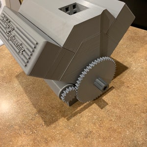以下が含まれることがあります： 側面に「Walgreen Engineering」という文字が書かれた灰色の3Dプリントされたオブジェクト。オブジェクトの下部にギアシステムがあります。
