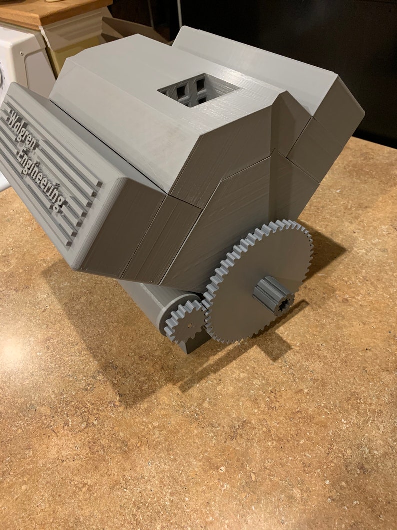 以下が含まれることがあります： 側面に「Walgreen Engineering」という文字が書かれた灰色の3Dプリントされたオブジェクト。オブジェクトの下部にギアシステムがあります。
