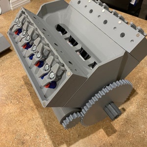 以下が含まれることがあります： 歯車システムを備えたグレーの3DプリントV8エンジンのモデル。エンジンは、ピストンとコンロッドが見える詳細な設計になっています。歯車システムはエンジンの底部に取り付けられており、大きな歯車と小さな歯車があります。