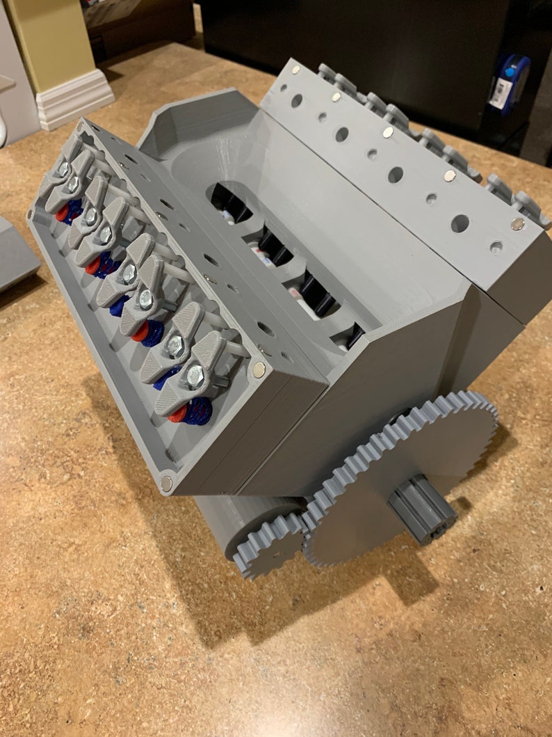 以下が含まれることがあります： 歯車システムを備えたグレーの3DプリントV8エンジンのモデル。エンジンは、ピストンとコンロッドが見える詳細な設計になっています。歯車システムはエンジンの底部に取り付けられており、大きな歯車と小さな歯車があります。