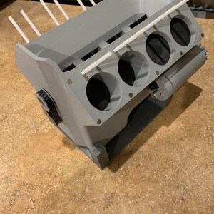 以下が含まれることがあります： 白いプラスチック製の棒がシリンダーに挿入された、グレーの3DプリントされたV8エンジンのモデル。このモデルは、ペイントブラシやその他のツールを保持するために設計されています。