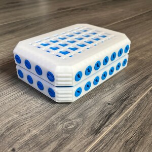 以下が含まれることがあります： 上部にグリッドパターン、縁に青い円がついた白いプラスチック製の箱。箱は閉まっており、2つの部分で構成されているように見える。