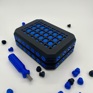 May include: A rectangular black storage case featuring a grid pattern on the top surface with blue inserts. The sides of the case are lined with blue circular accents. A small blue handled screwdriver with a metal tip is positioned nearby.