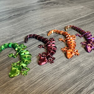 以下が含まれることがあります： プラスチック製のカラフルなヤモリの置物4つ。ヤモリは緑、紫、オレンジ、赤の色合いです。尻尾を丸めて這っているポーズをしています。