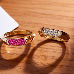 Peut inclure: Deux bagues en or, l'une avec un sertissage de pierres roses et l'autre avec des pierres claires. Une boîte cadeau blanche avec un ruban marron et une carte avec l'inscription "With Love" sont également visibles.