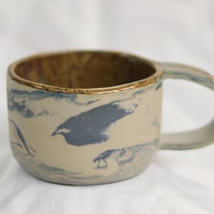 May include: A ceramic mug with a distinctive design. The mug has a beige exterior with blue and grey abstract patterns. The interior is a warm brown colour. The handle is a matching beige. A handmade, artistic piece.