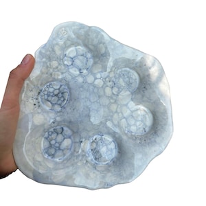 May include: A handmade, organic-shaped ceramic serving dish in shades of white and blue. The dish has five circular indentations and a textured surface. The dish is held by a hand against a white background.