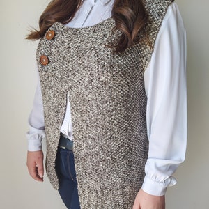 Op de afbeelding: Een bruin en wit gebreid vest met een lange, open voorkant en twee bruine knopen. Het vest wordt gedragen over een wit overhemd met knopen en een blauwe jeans.