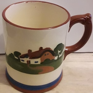 Może przedstawiać: Biały kubek ceramiczny z brązowym uchwytem. Kubek ma namalowany obrazek przedstawiający brązowo-biały domek z czerwonym dachem, otoczony zielonymi drzewami i brązową ścieżką.