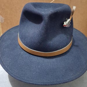 May include: A navy blue fedora hat with a wide brim and a light brown leather band. The hat has a decorative pin and a feather. This hat is a stylish accessory.