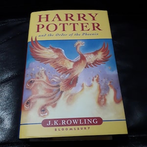 Puede incluir: Un libro titulado "Harry Potter y la Orden del Fénix" de J.K. Rowling. La portada presenta un fénix en tonos rojos y naranjas sobre un fondo azul. También se ve el nombre de la editorial, Bloomsbury.