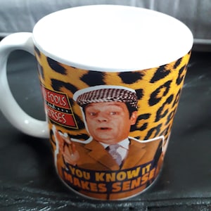 May include: White ceramic mug with a handle. The mug features a leopard print design and a graphic of a man in a brown suit and cap. Text on the mug reads "Fools and Horses" and "You know it makes sense."