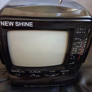 May include: A black New Shine mini personal television with an AM/FM radio. The device features a screen, a side-mounted frequency tuning panel with numbers, a volume dial, an earphone jack, and switches for radio, TV, and band selection.