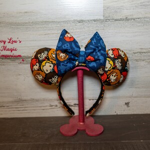 May include: A pair of blue and white Minnie Mouse ears with a bow. The ears are covered in a fabric with a pattern of Harry Potter characters. The bow is blue with white musical notes.