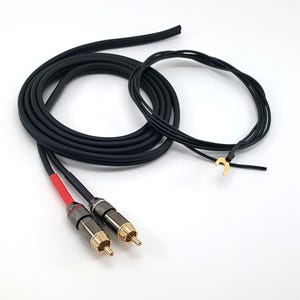 Könnte beinhalten: Schwarze Audiokabel mit goldfarbenen Steckern und roter Schrumpfschlauchisolierung. Die Kabel sind auf einer weißen Oberfläche aufgerollt. Ein separates schwarzes Kabel mit einem goldfarbenen Stecker ist ebenfalls vorhanden.