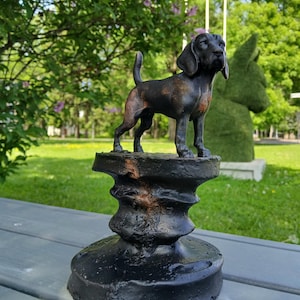 Könnte beinhalten: Eine schwarz-braune Metallhundestatue auf einem dekorativen Sockel. Der Hund hat Schlappohren und einen kurzen Schwanz. Die Statue steht vor einem verschwommenen Hintergrund aus grünem Gras und Bäumen.