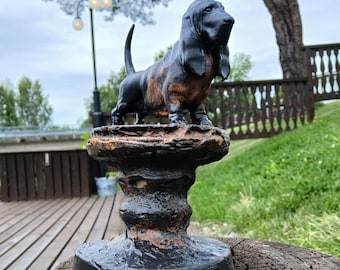 May include: A black and rust-colored cast iron Basset Hound figurine standing on a textured, cylindrical pedestal base. The dog has long, drooping ears and a short, upright tail, with a weathered finish showing metallic highlights.