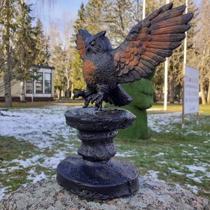 Könnte beinhalten: Eine schwarze Metall-Eulenstatue mit orangefarbenen Akzenten, die auf einem schwarzen Metallsockel thront. Die Eule befindet sich in einer ausgebreiteten Flügelhaltung mit dem Kopf zur Seite gedreht. Die Statue steht im Freien auf einer Steinoberfläche.