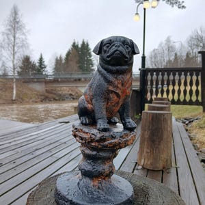 Puede incluir: Una estatua de un pug negro y óxido se encuentra sobre un pedestal decorativo. El pug está posado sobre una base desgastada y texturizada, con una cara detallada y expresiva. La estatua está colocada sobre una superficie de madera.