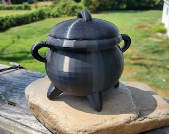 May include: A small black cauldron with a lid, sitting on a light brown rock. The cauldron has three legs and a handle on the lid.