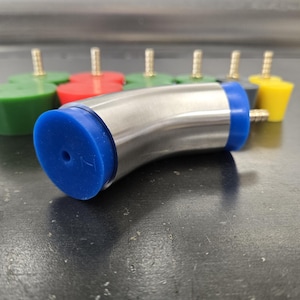 May include: A stainless steel curved pipe with blue rubber ends and a small hole. Several colorful rubber stoppers with brass fittings are in the background. The stoppers are green, red, and yellow.