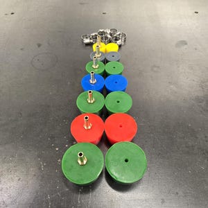 May include: An assortment of colorful rubber grommets with brass fittings, arranged in a vertical line. The grommets are in various colors, including green, red, blue, yellow, and gray. A collection of metal clamps is at the top.
